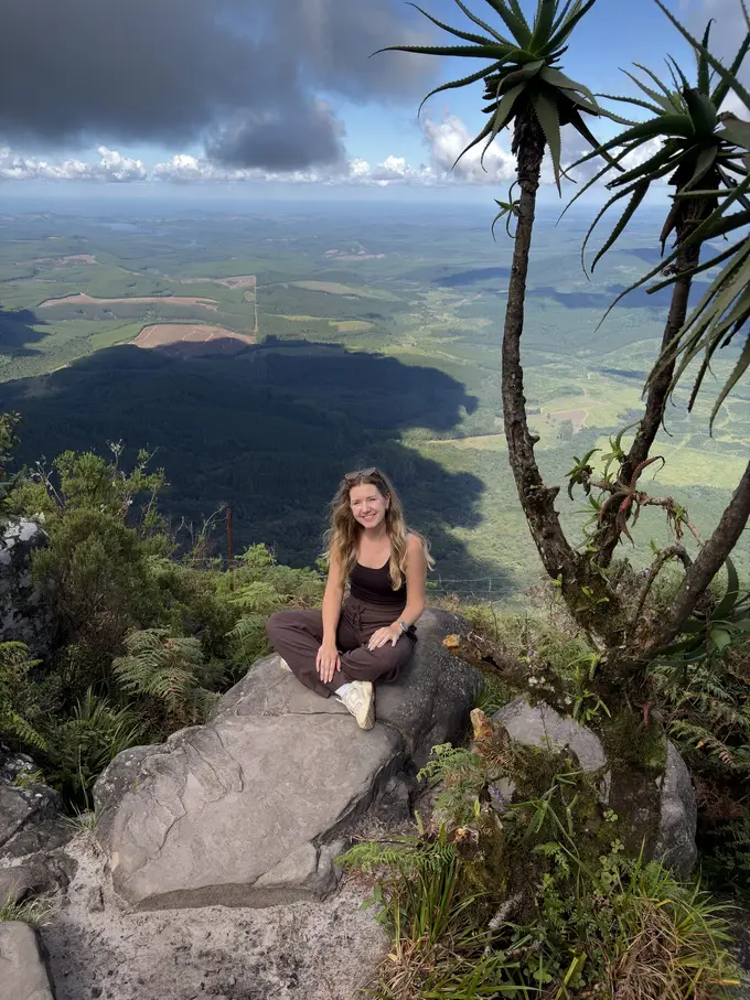 Rebecca sitzt auf einem Felsen neben hohen Aloe-Pflanzen und blickt über ein weites grünes Tal