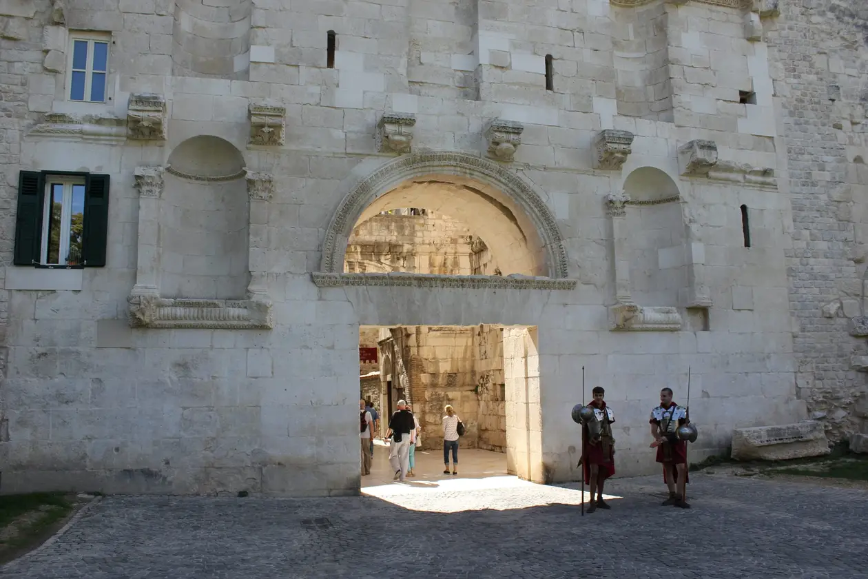 Ein Tor in der hellen Außenmauer des Diokletianpalasts mit Rundbogen und Nischen, Besucher gehen hindurch, daneben zwei Männer in römischen Soldatenkostümen