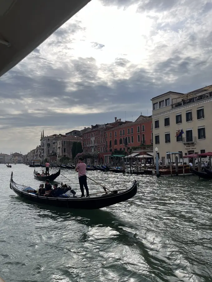Eine Gondel mit Gondoliere im gestreiften Hemd auf dem Canal Grande, am rechten Ufer Hotels mit Anlegern und Sonnenschirmen, darüber ein bewölkter Himmel