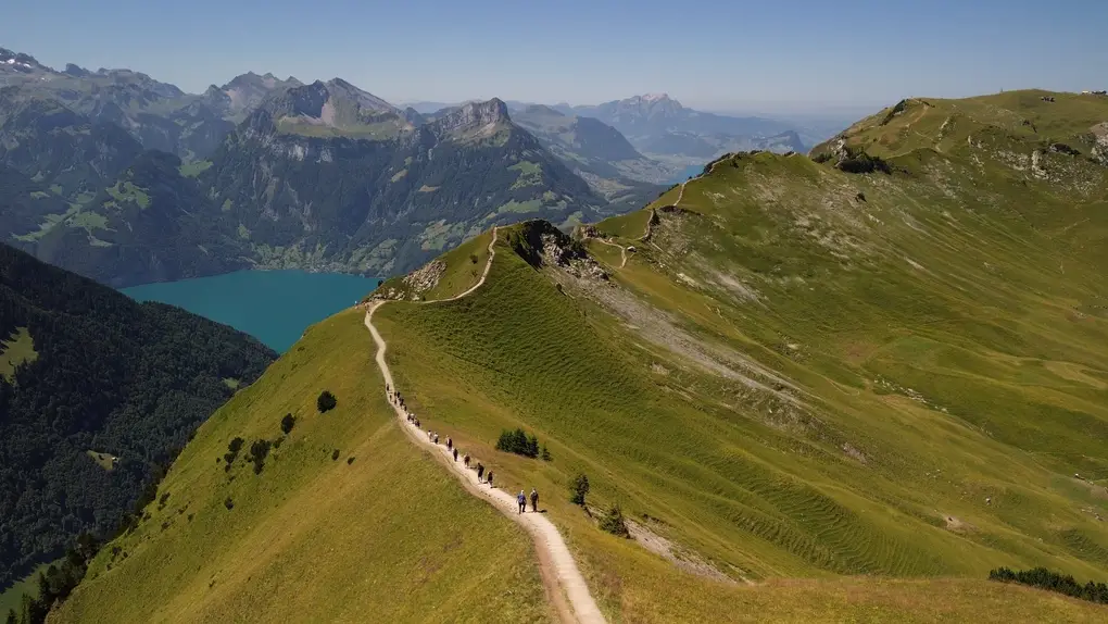 Drohnenfoto über einen grasbewachsenen Grat, auf dem ein schmaler Pfad mit einzelnen Wanderern entlangführt, links unten ein türkiser See