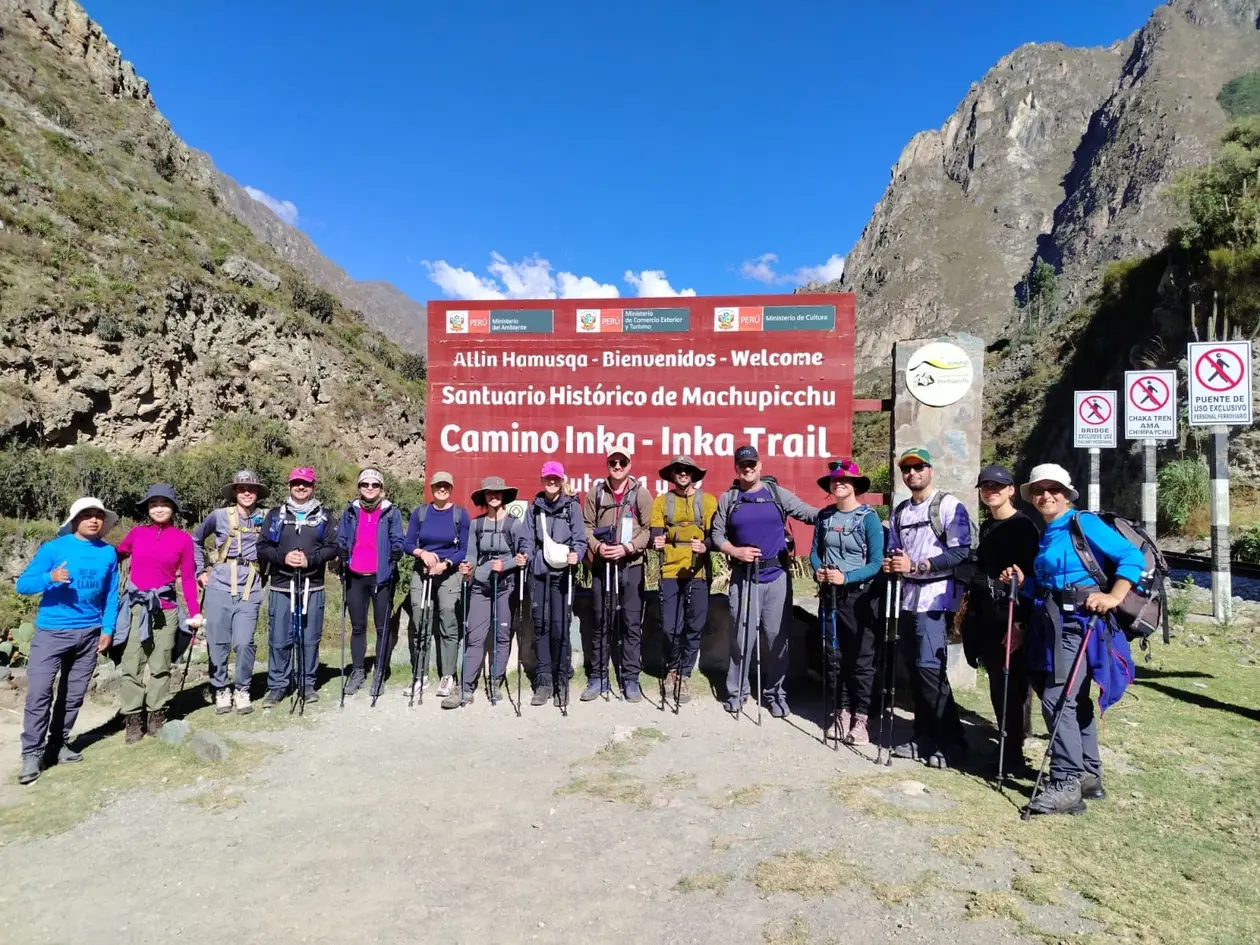 Die Wandergruppe mit Wanderstöcken in einer Reihe vor dem roten Schild „Camino Inka – Inka Trail“, dahinter steile Felswände unter blauem Himmel