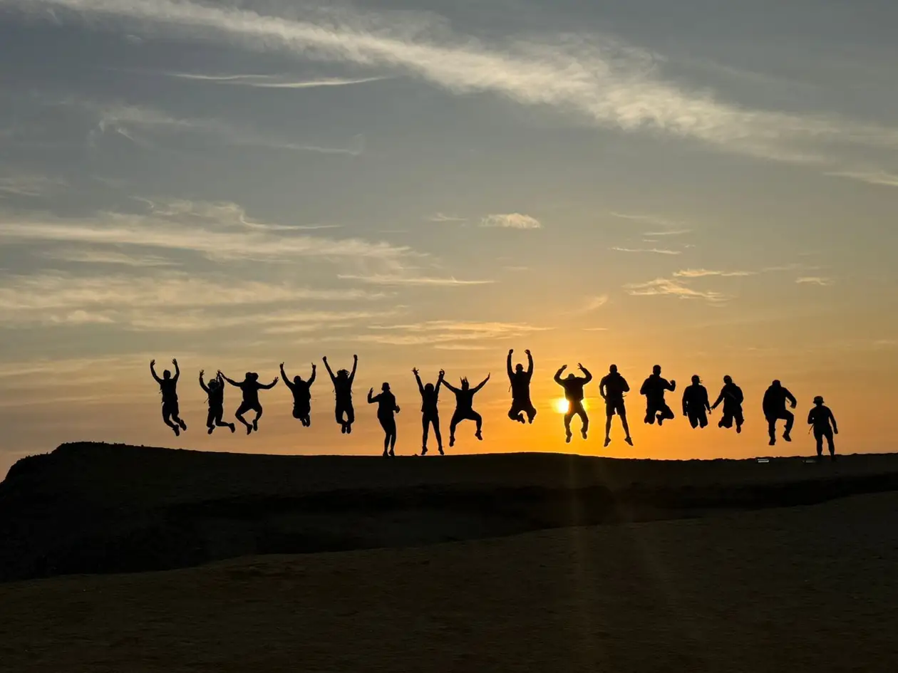 Eine Wandergruppe springt als Silhouette auf einem Felsrücken vor der tief stehenden orangen Sonne
