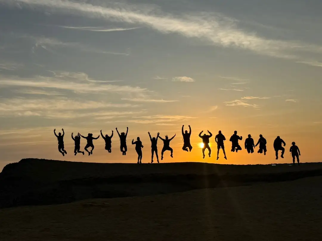 Eine Wandergruppe springt als Silhouette auf einem Felsrücken vor der tief stehenden orangen Sonne