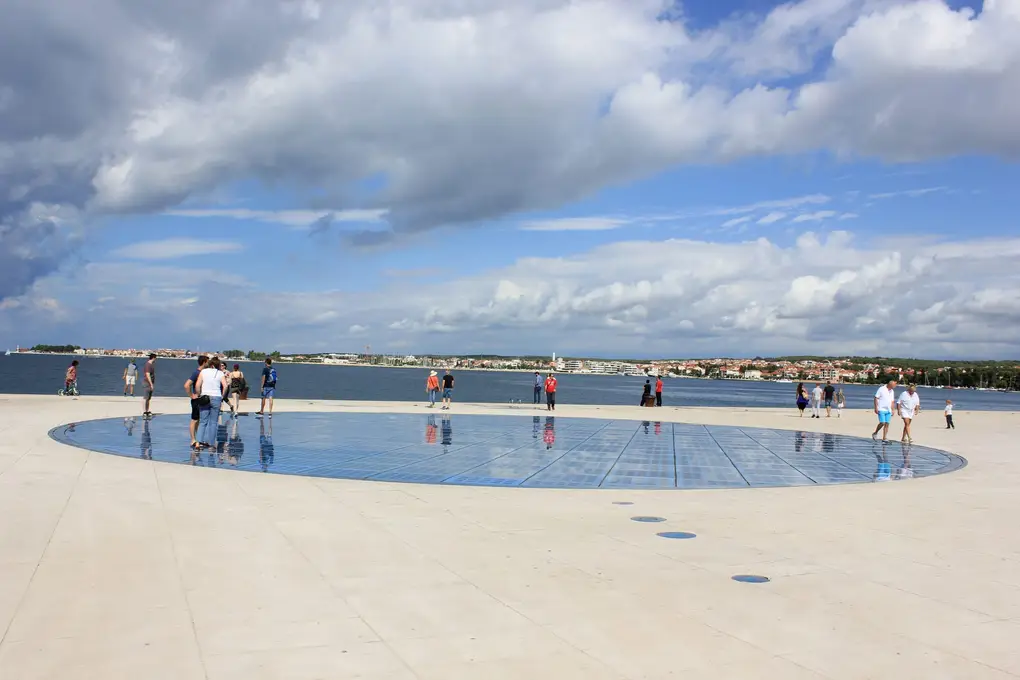 Eine große runde Glasfläche im Steinboden am Ufer, in der sich Menschen spiegeln, dahinter das Meer und die Küste gegenüber