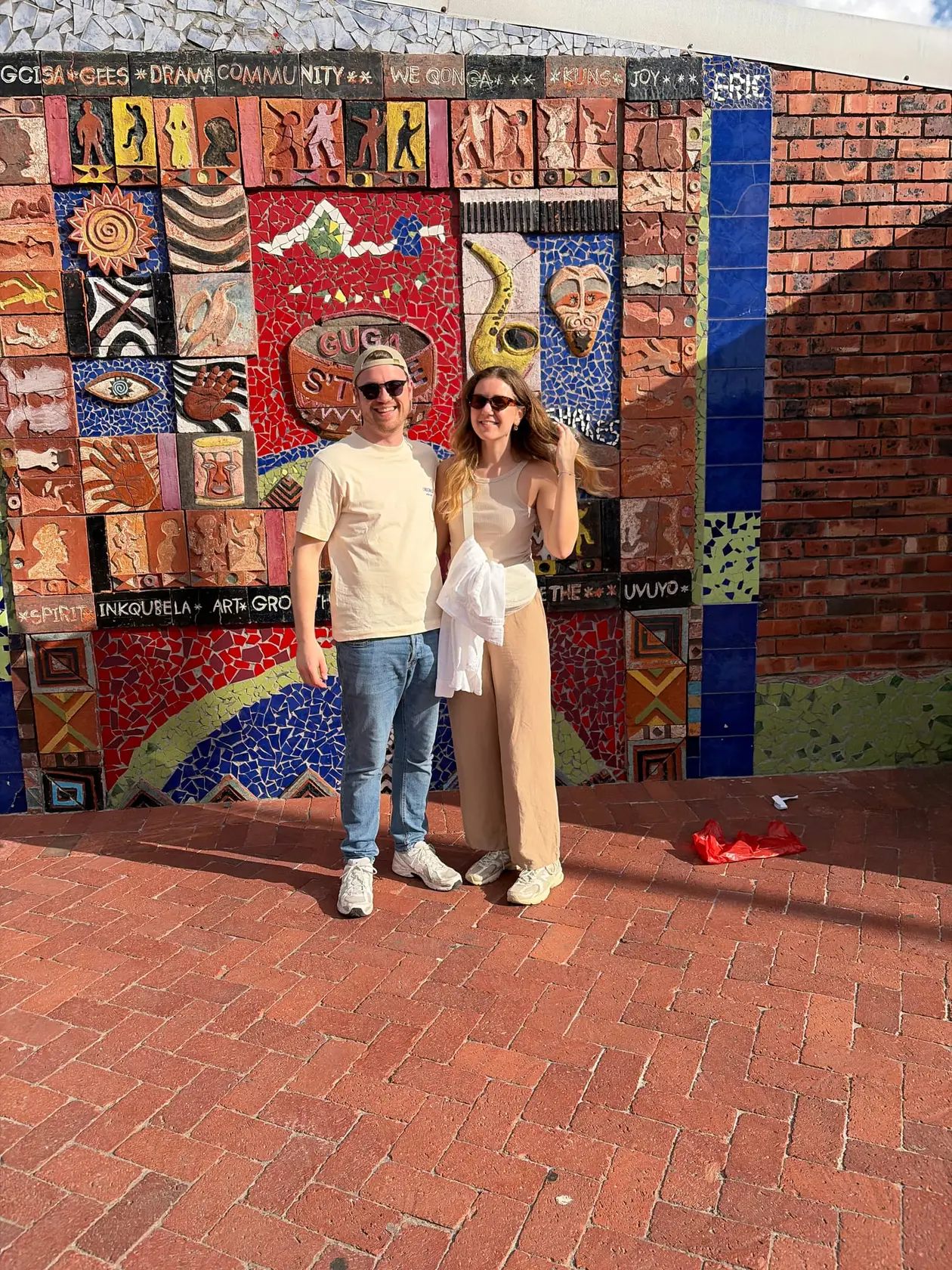 Max und Rebecca stehen vor einer großen bunten Mosaikwand mit afrikanischen Mustern und der Aufschrift „Guga S'thebe“