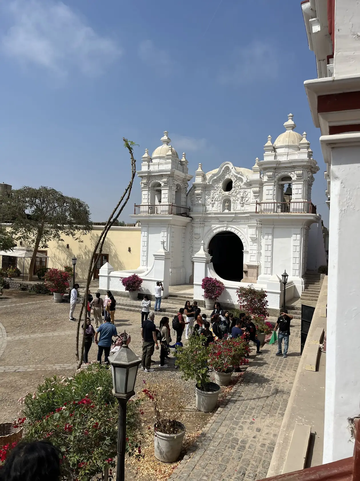 Weiße Barockkapelle mit zwei Glockentürmen an einem gepflasterten Hof mit Blumenkübeln, davor eine Reisegruppe