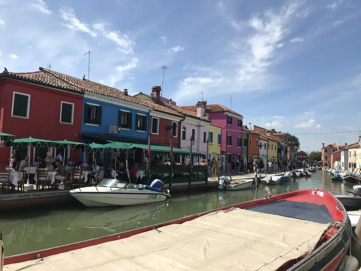 Häuserzeile in Rot, Blau, Gelb und Rosa an einem Kanal, davor Restaurantschirme und festgemachte Motorboote