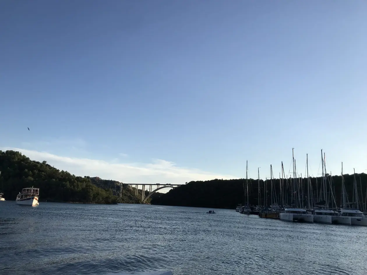 Segelboote und Katamarane an einem Hafenkai, links ein Ausflugsboot, im Hintergrund eine Bogenbrücke über den Fluss