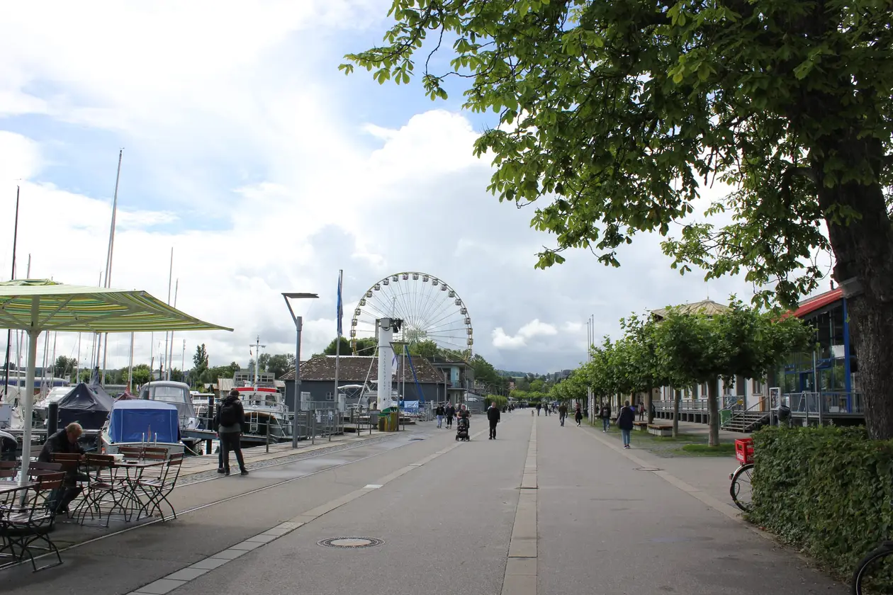 Breite Promenade am Hafen mit Spaziergängern, links Boote und Masten, am Ende der Promenade ein Riesenrad