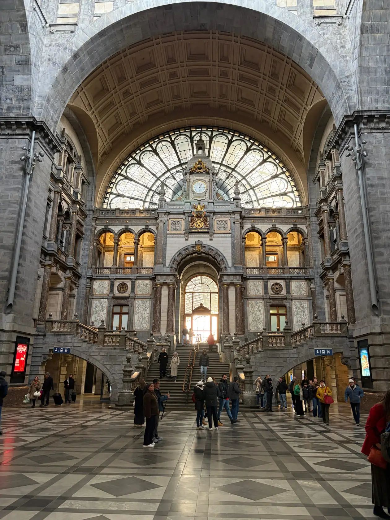 Die Eingangshalle von Antwerpen-Centraal mit Steinbogen, großem Rundfenster, Uhr und doppelter Treppe, auf dem gemusterten Boden Reisende