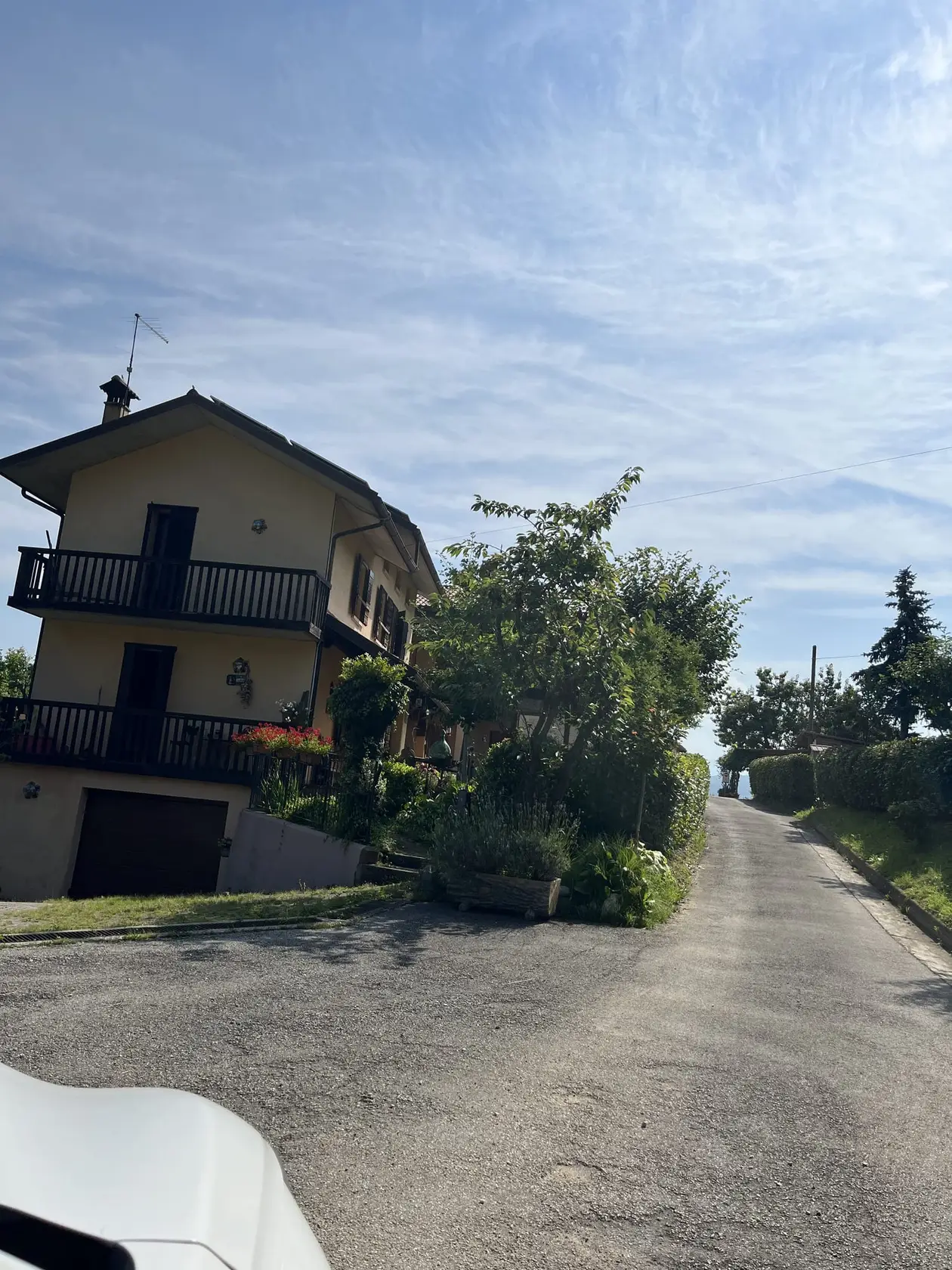Ein zweistöckiges Haus mit dunklen Holzbalkonen und roten Blumen an einer schmalen asphaltierten Straße, die rechts steil ansteigt, vorne links die Motorhaube eines weißen Autos