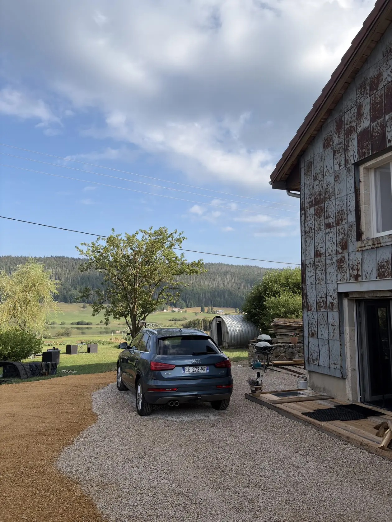 Ein Haus mit fleckiger, verwitterter Fassadenverkleidung, davor ein Kiesplatz mit einem geparkten Auto, im Hintergrund Wiesen und Wald