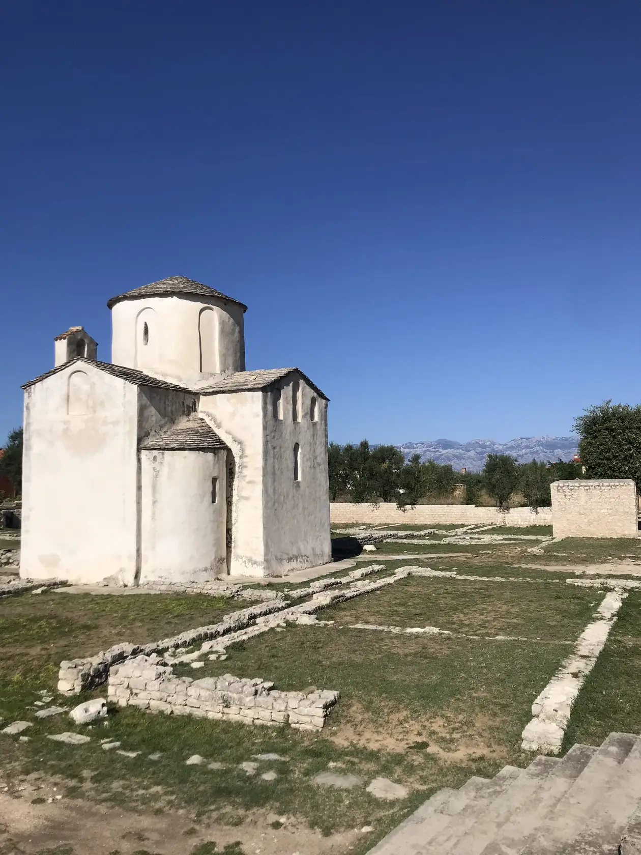 Eine kleine weiße Steinkirche mit Kuppel auf einer Wiese, davor Grundmauern im Gras, dahinter ein Gebirgszug