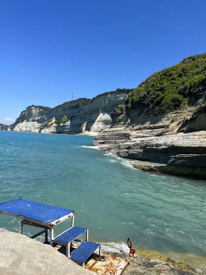 Helle Steilküste über türkisem Wasser, vorne eine blaue Badeplattform mit Stufen auf dem Fels