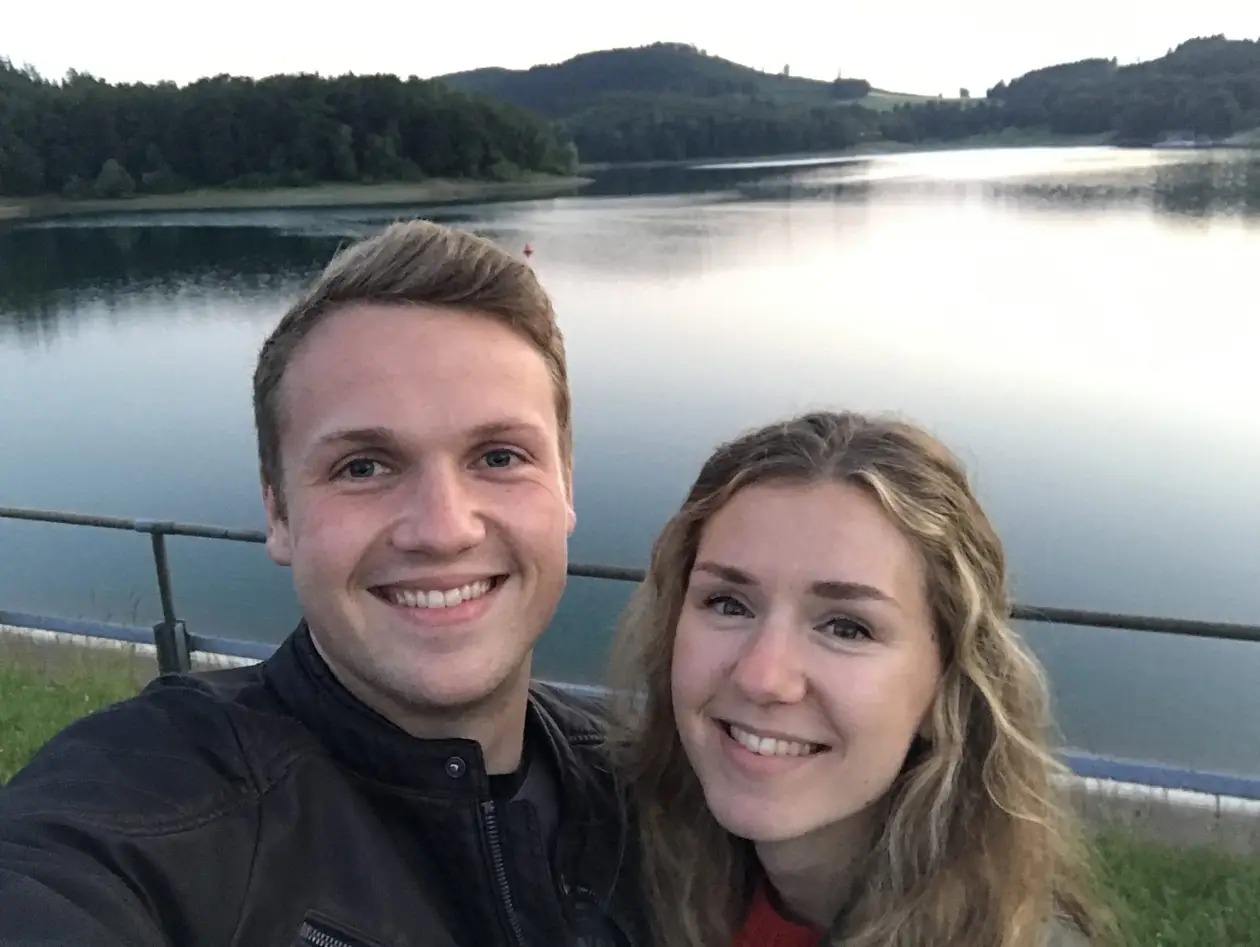 Selfie von Max und Rebecca am Geländer über dem Hennesee in der Dämmerung, hinter ihnen der glatte See und bewaldete Hügel