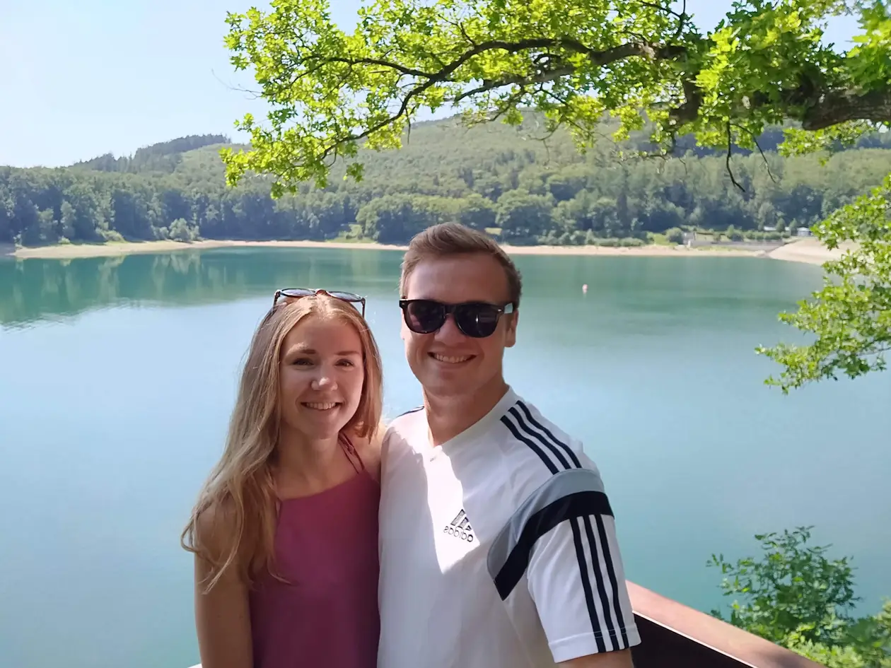 Rebecca und Max stehen mit Sonnenbrillen im Haar und auf der Nase an einem Geländer, hinter ihnen der türkisfarbene Hennesee und bewaldete Hänge, oben ragt ein Eichenast ins Bild