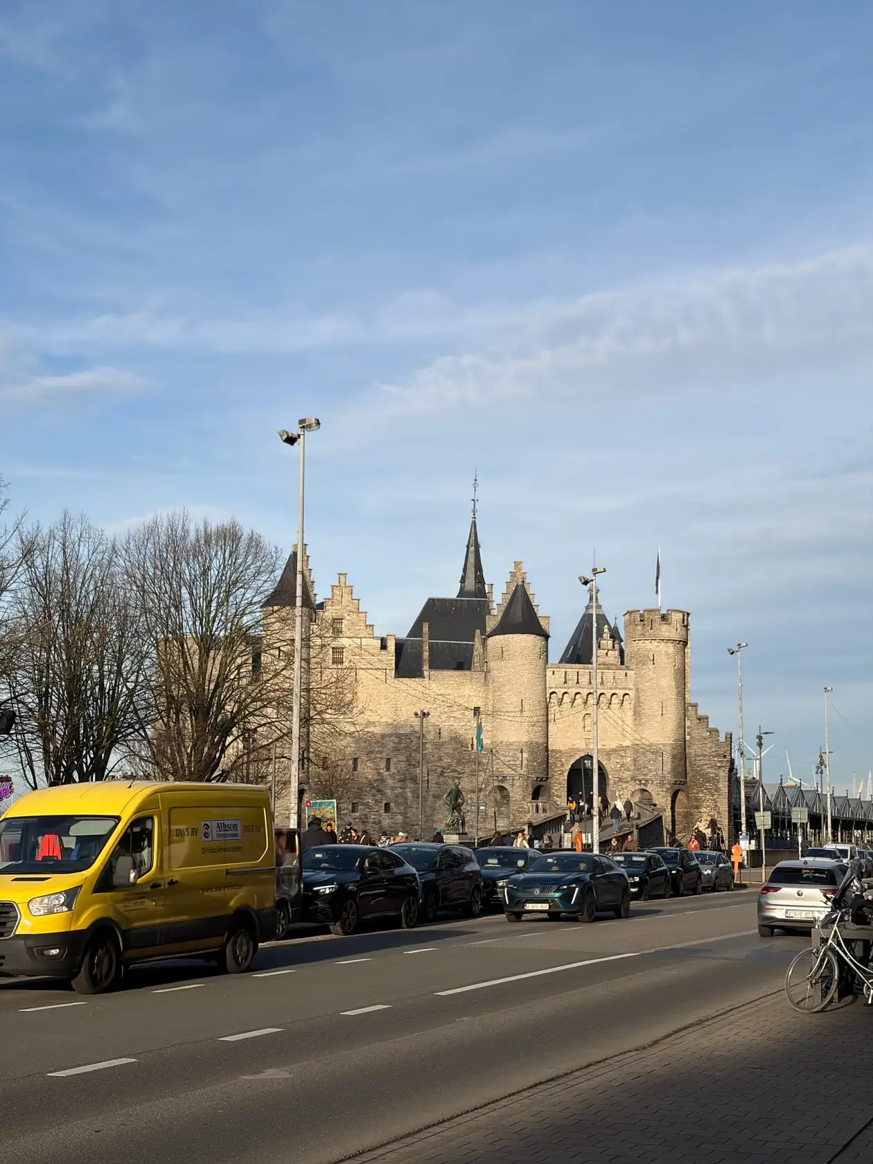 Die Burg Het Steen aus hellem Stein mit Rundtürmen und spitzen Dächern in der tief stehenden Sonne, davor eine Straße mit Autos und einem gelben Lieferwagen
