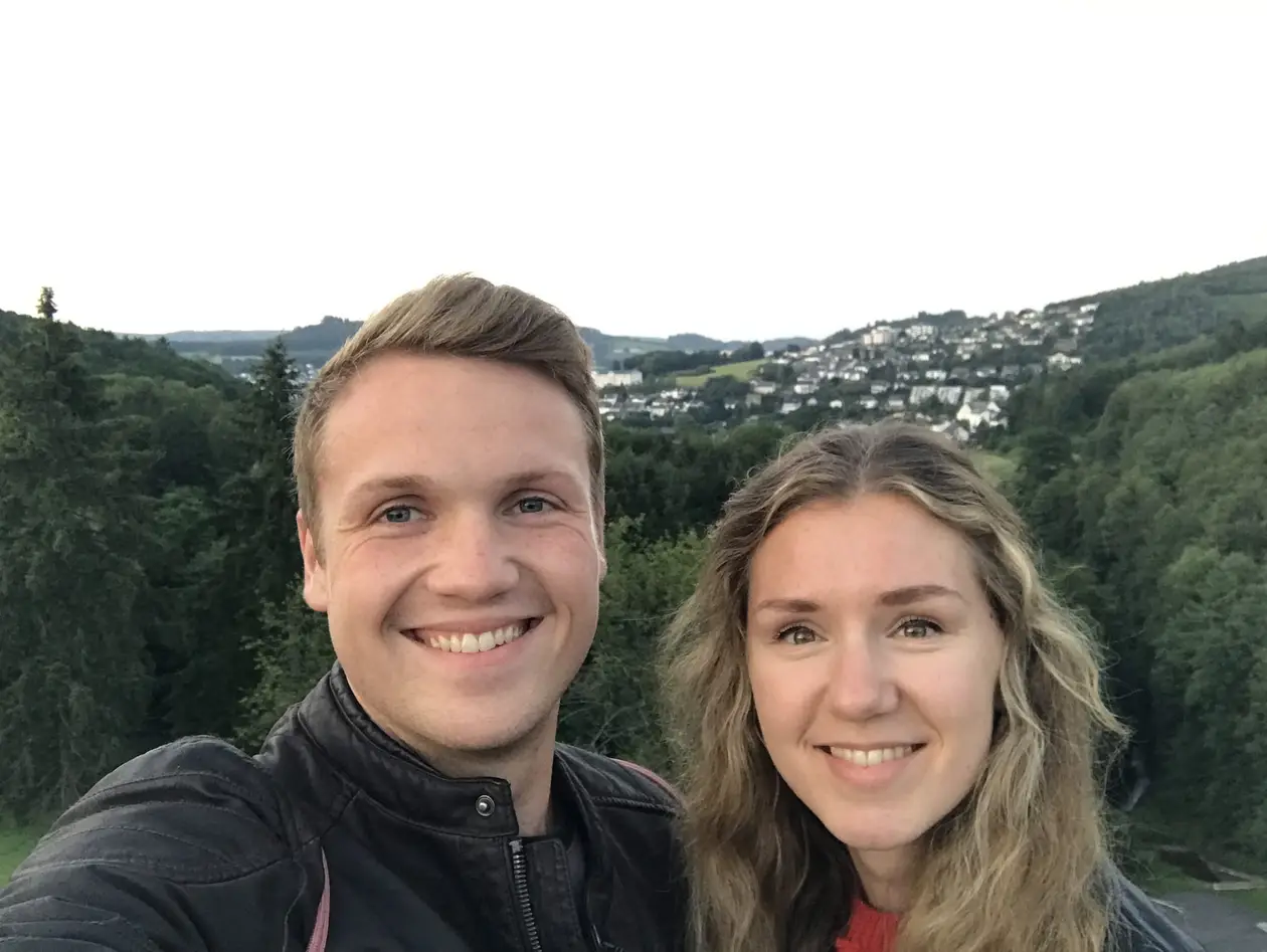 Selfie von Max und Rebecca auf einer Aussichtsplattform, hinter ihnen ein bewaldetes Tal und eine Ortschaft am Hang