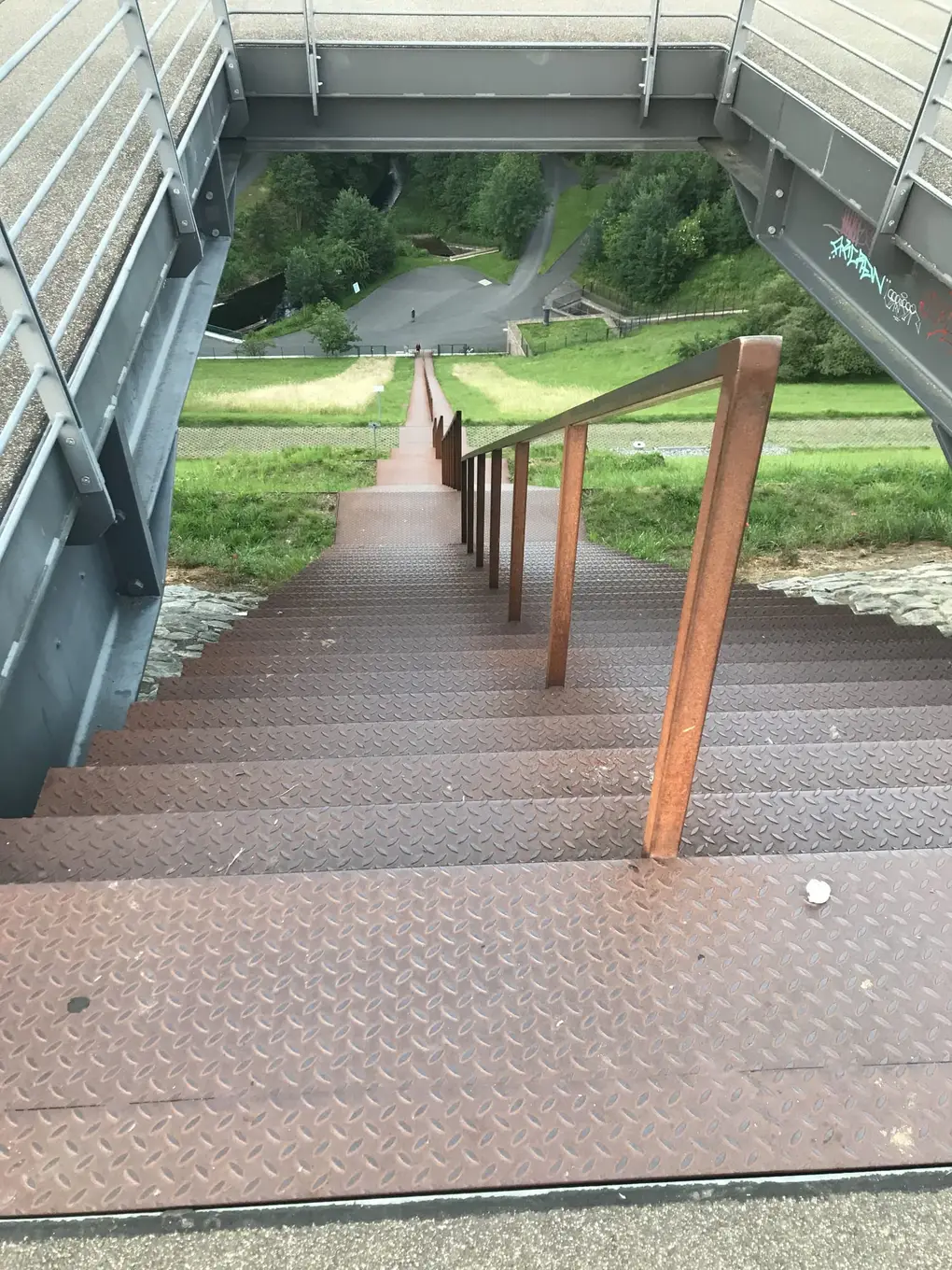 Blick von oben eine lange rostbraune Stahltreppe mit Handlauf hinunter, die über eine Grasböschung bis zu einem Weg im Tal führt, links und rechts die grauen Stahlträger einer Plattform
