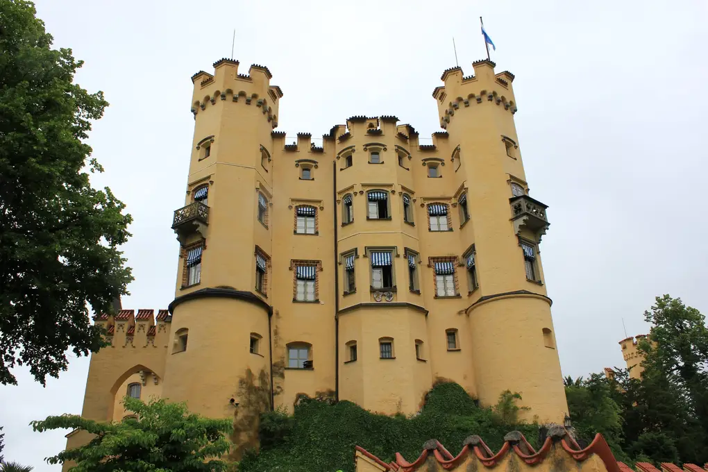 Schloss Hohenschwangau von unten, ein gelbes Schloss mit Zinnen, zwei Ecktürmen und blau-weiß gestreiften Markisen, oben eine blau-weiße Fahne
