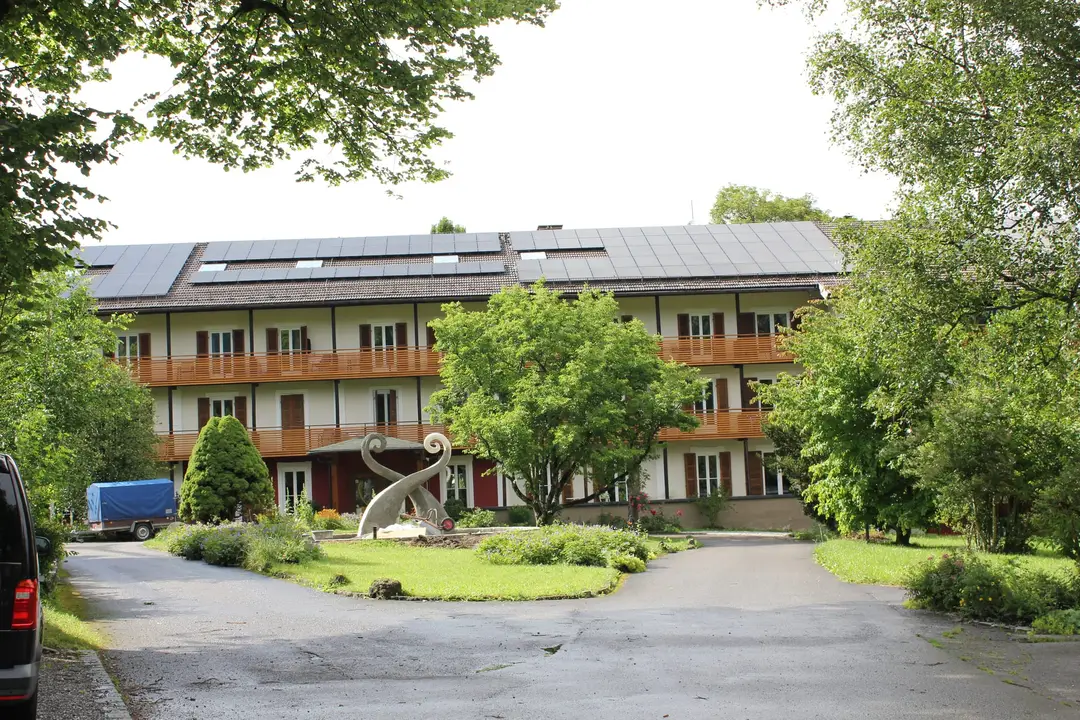 Ein langes, helles Haus mit Holzbalkonen und Solarmodulen auf dem ganzen Dach, davor eine Einfahrt, ein Rondell mit Blumen und eine geschwungene Skulptur