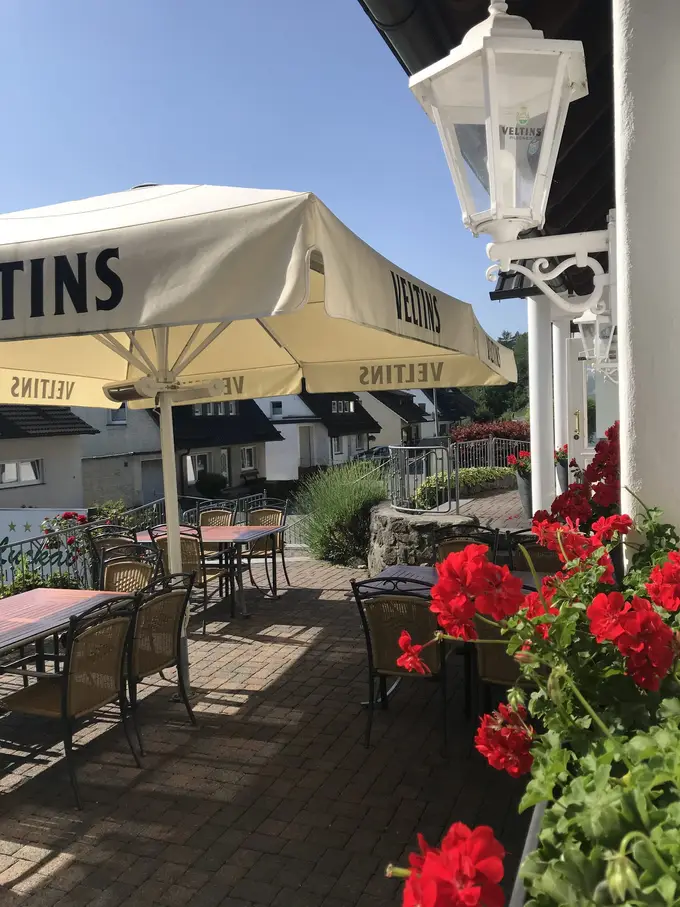 Leere Terrasse mit Holztischen, Korbstühlen und einem großen Sonnenschirm, rechts eine weiße Laterne an der Hauswand und rote Geranien im Blumenkasten