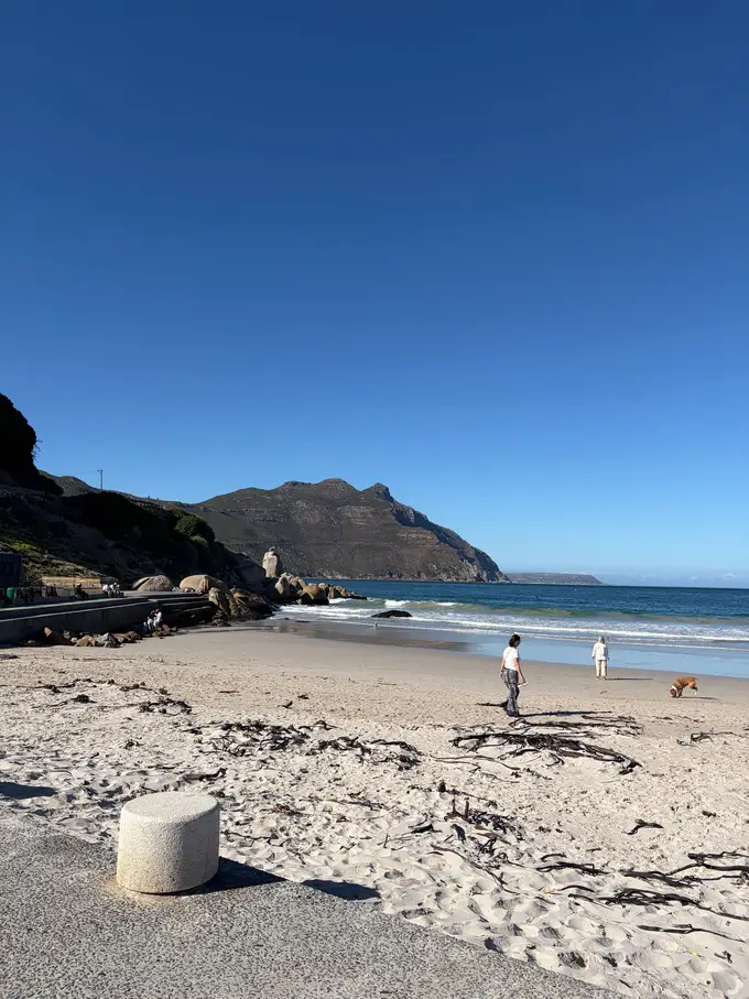 Sandstrand der Hout Bay mit Bergen im Hintergrund, am Ufer zwei Spaziergänger mit Hund