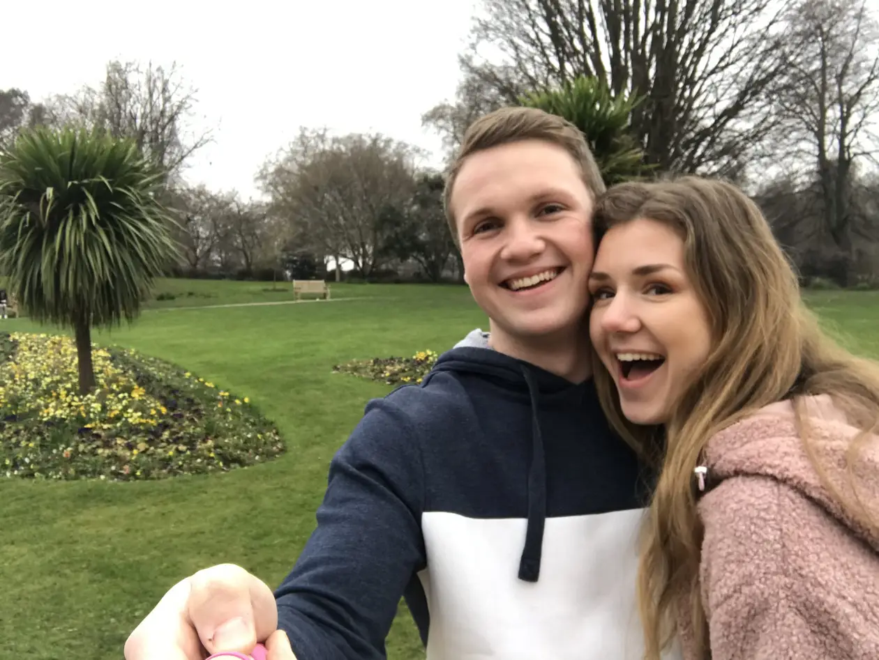 Max und Rebecca machen im Hyde Park ein Selfie mit dem Selfiestick, hinter ihnen Rasen, Blumenbeete und eine Palme