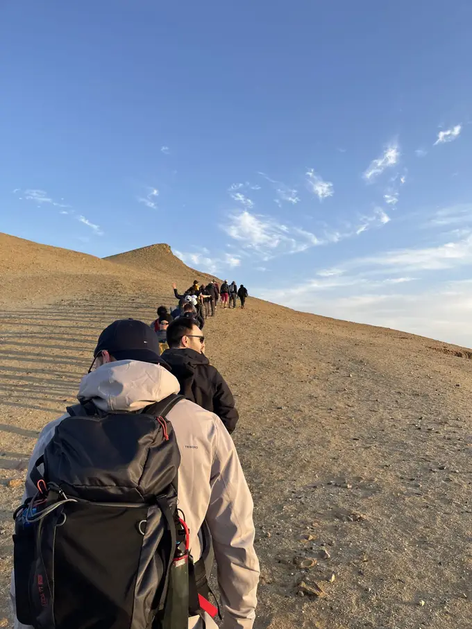 Wandernde laufen hintereinander einen Sandkamm hinauf, im Vordergrund ein Mann mit Rucksack von hinten