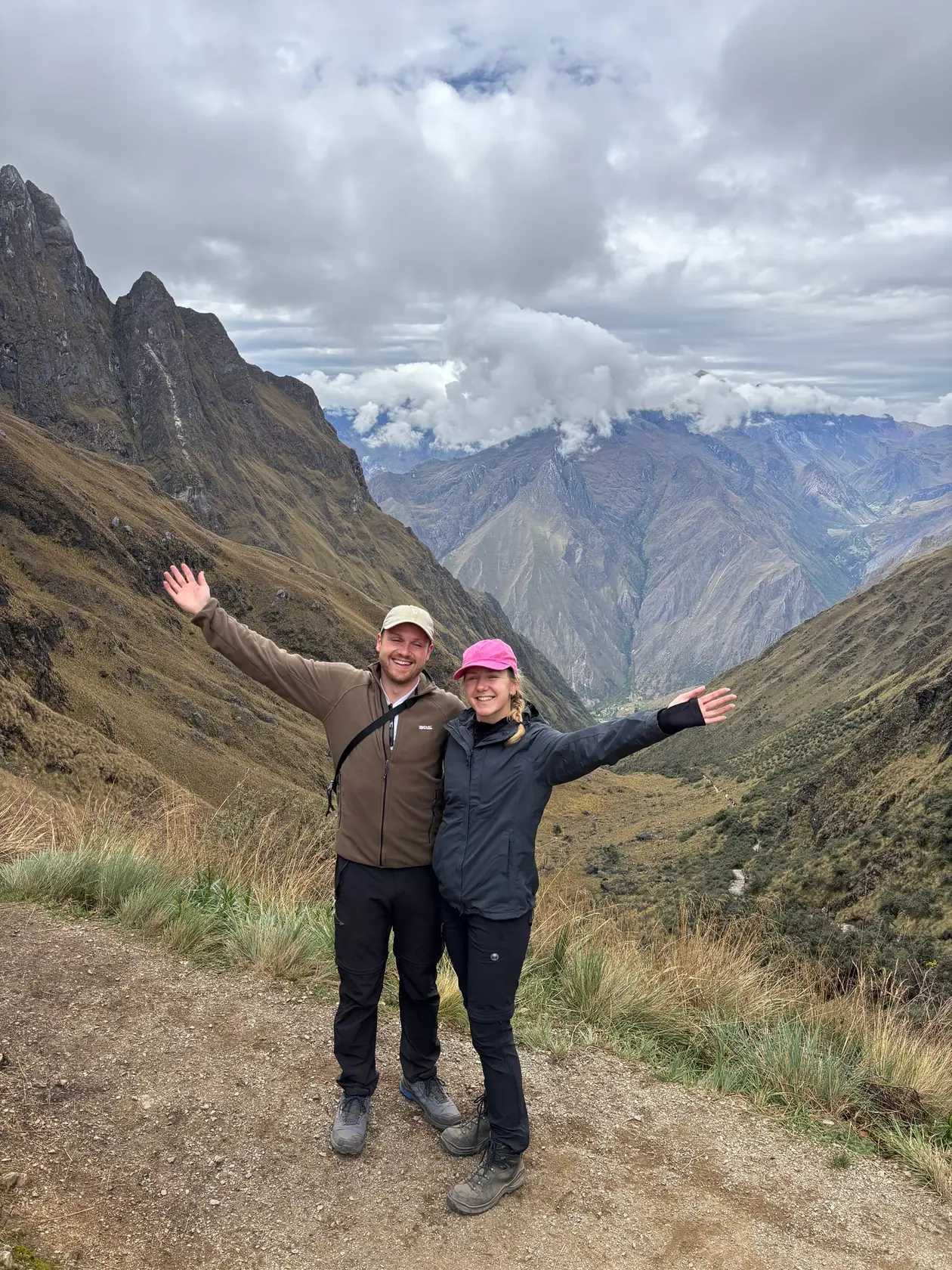 Max und Rebecca mit ausgebreiteten Armen auf einem Bergweg, hinter ihnen ein tiefes grünes Tal und Wolken über den Gipfeln