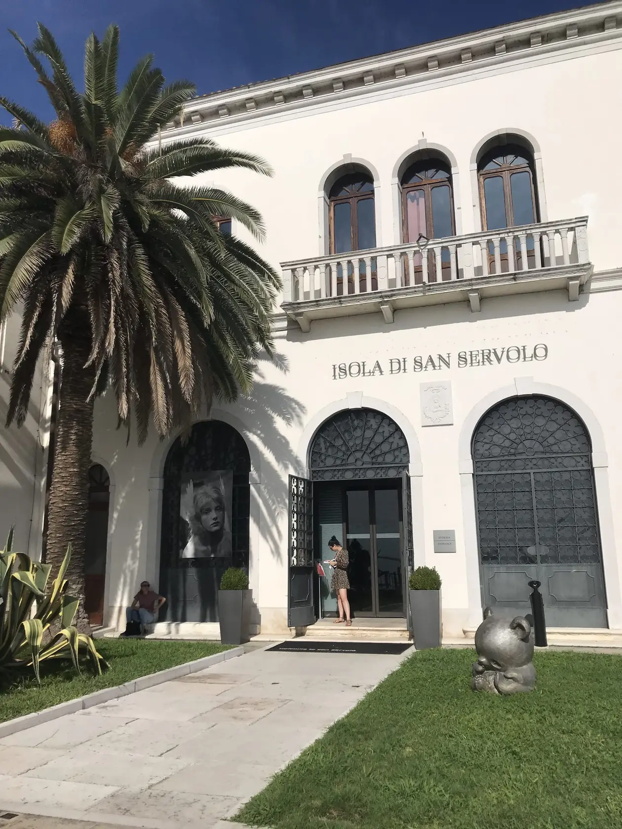 Weißes Gebäude mit Bogenfenstern, Balkon und der Aufschrift Isola di San Servolo, davor eine Palme und Rasen