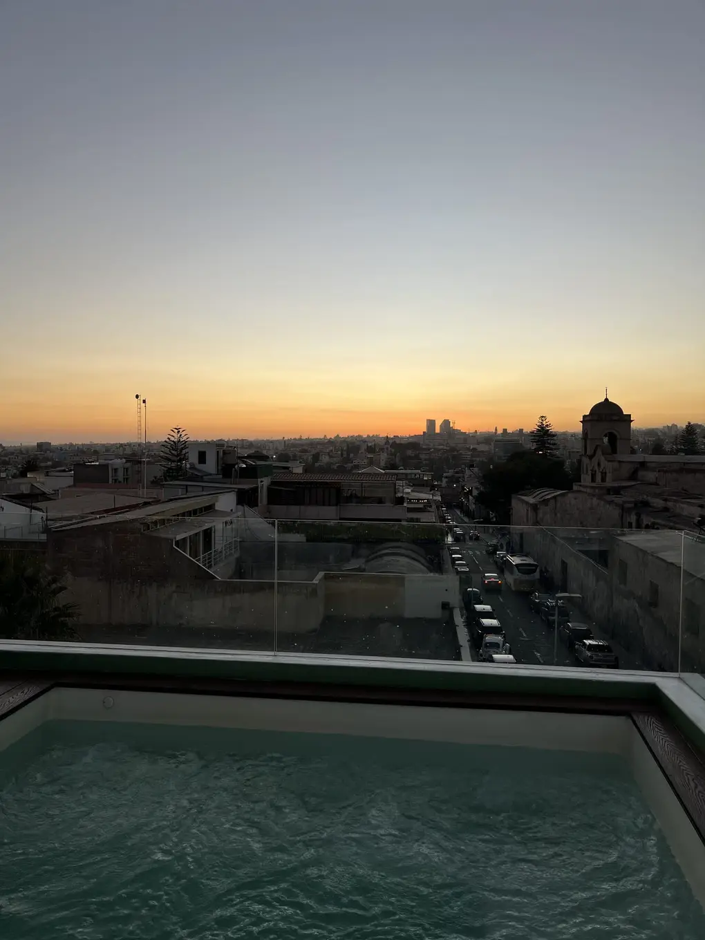 Jacuzzi am Rand der Dachterrasse, dahinter die Dächer der Stadt und ein Kirchturm im Sonnenuntergang
