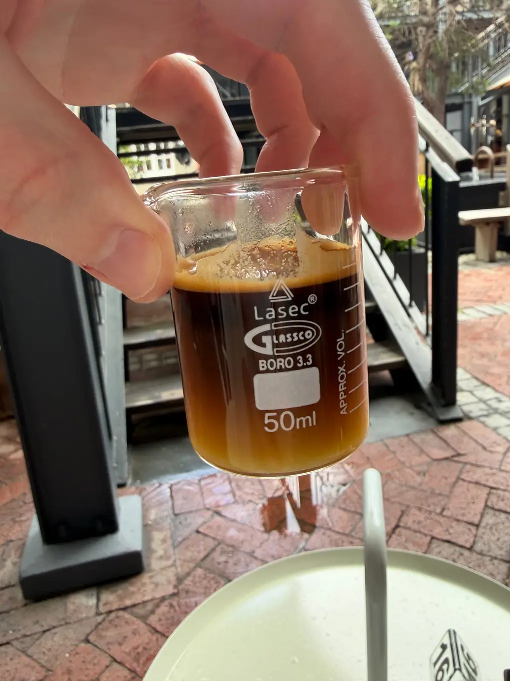 Eine Hand hält ein kleines Laborglas mit schwarzem Kaffee zwischen zwei Fingern