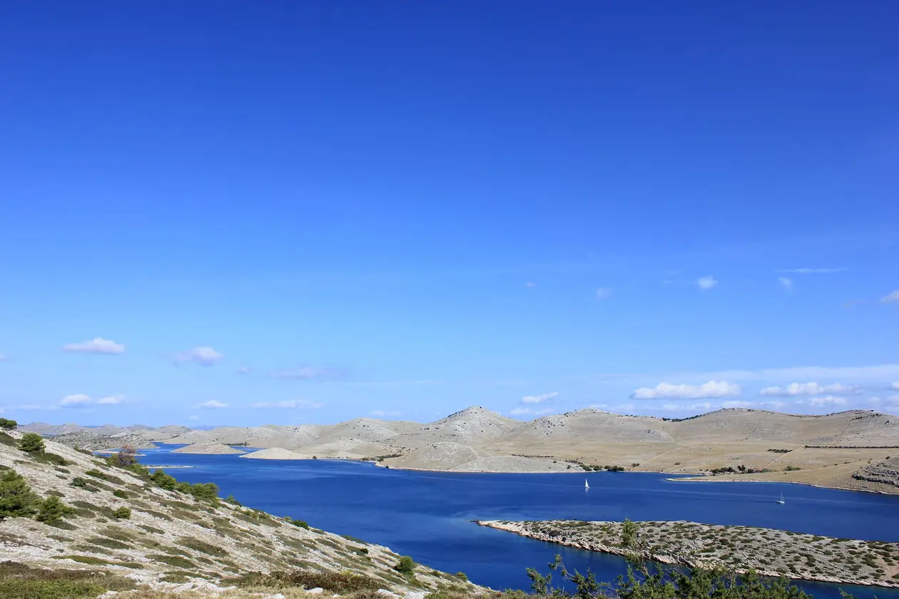 Blick von einer Anhöhe auf eine tiefblaue Meeresstraße zwischen hellen, kahlen Inseln, darauf zwei Segelboote