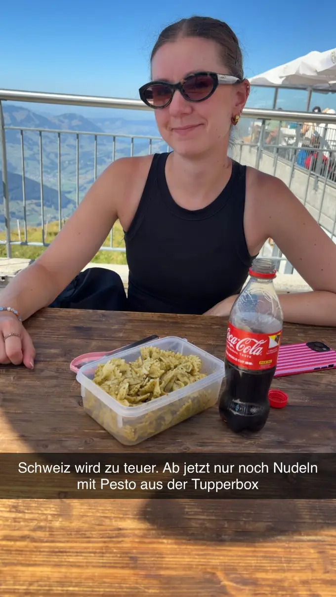 Rebecca sitzt an einem Holztisch vor einer Plastikdose mit Nudeln in Pesto und einer Flasche Cola, darüber eine eingeblendete Zeile: Schweiz wird zu teuer. Ab jetzt nur noch Nudeln mit Pesto aus der Tupperbox