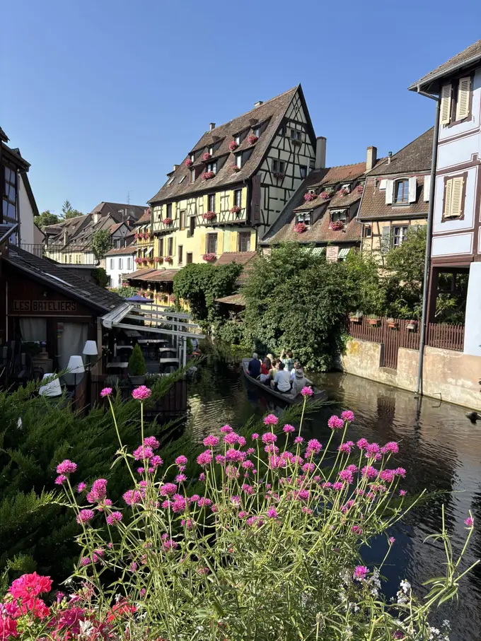 Ein flaches Boot mit Besuchern auf einem schmalen Kanal zwischen Fachwerkhäusern mit Blumenkästen, vorne pinke Blumen