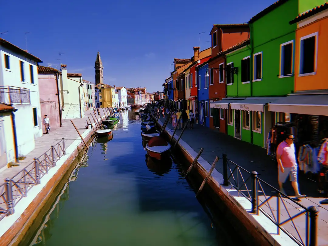 Kanal zwischen grünen, roten, orangen und rosa Häusern, im Hintergrund ein schiefer Glockenturm
