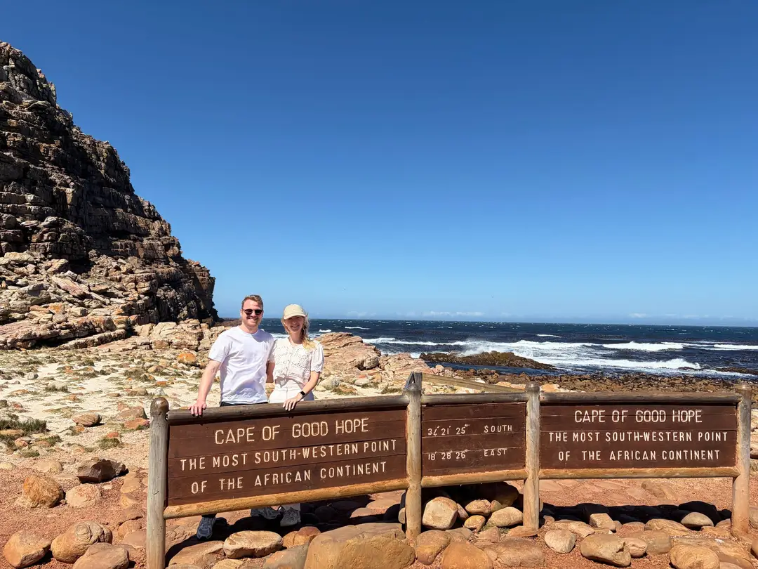 Max und Rebecca stehen vor dem braunen Schild „Cape of Good Hope – The Most South-Western Point of the African Continent“, dahinter Klippen und der Atlantik