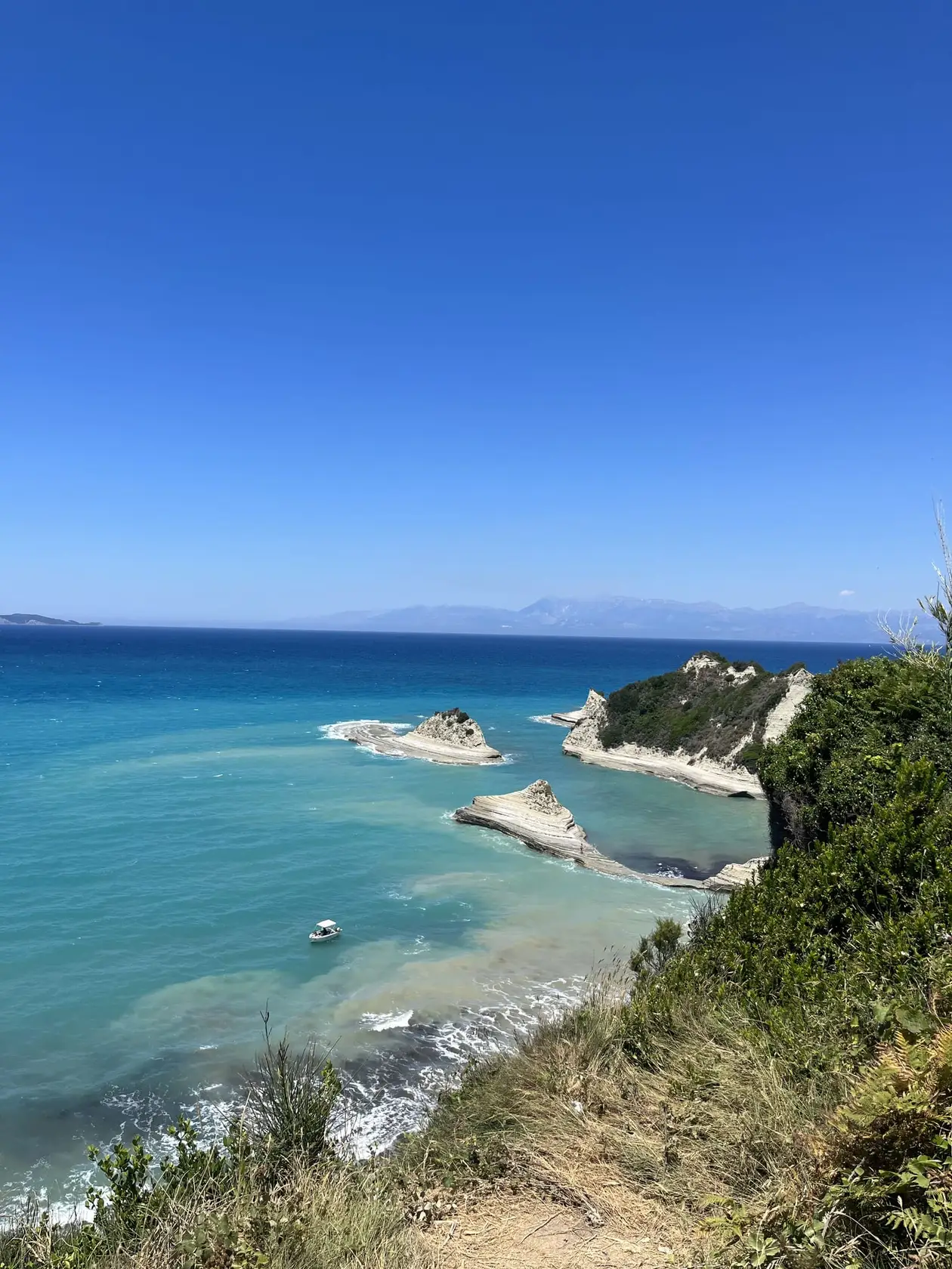 Blick von einem bewachsenen Hang hinunter auf weiße Felsen und Felsinseln in türkisem Wasser