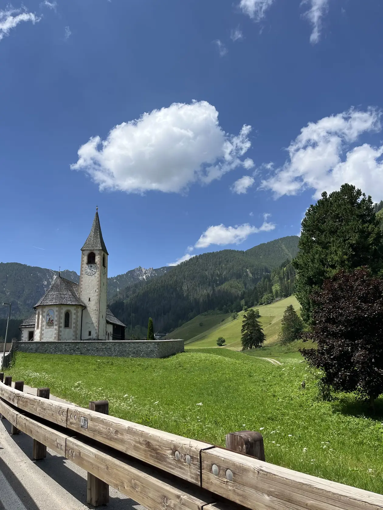 Eine weiße Dorfkirche mit spitzem Turm und Uhr hinter einer Friedhofsmauer auf einer grünen Wiese, dahinter bewaldete Berge, vorne ein hölzernes Straßengeländer