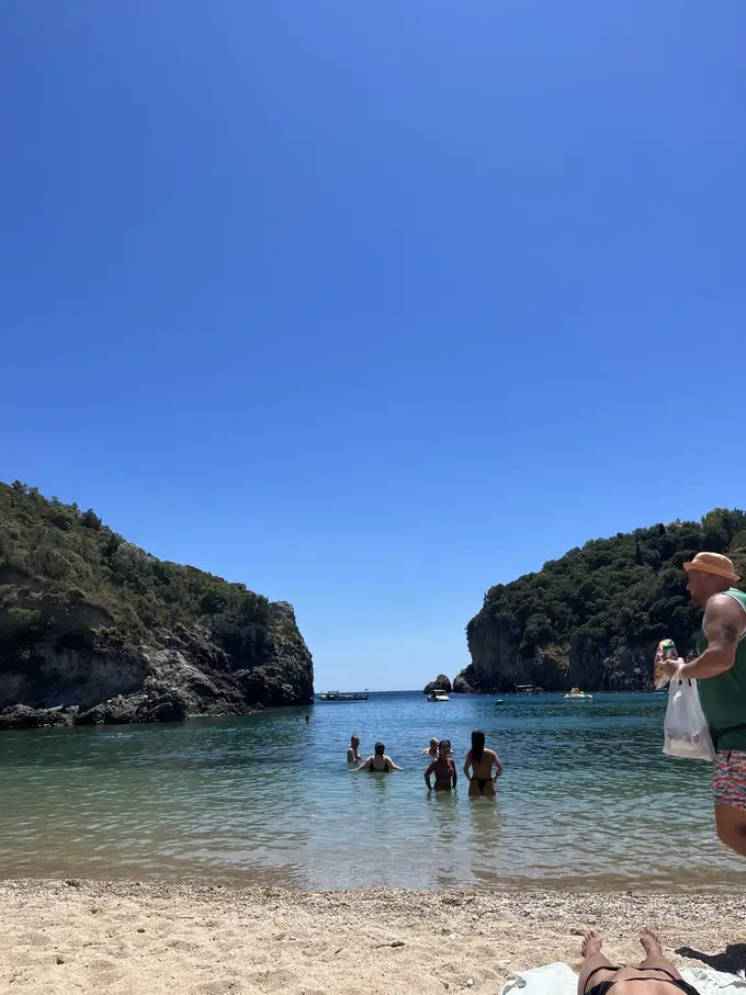 Kleiner Strand zwischen zwei bewaldeten Felsen, im flachen Wasser stehen Badende