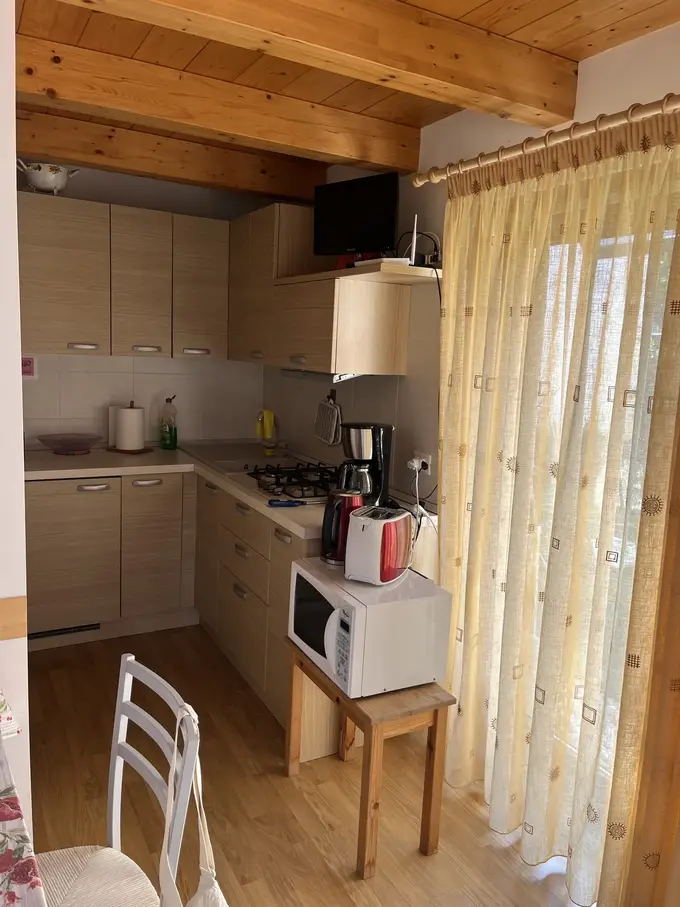 Kleine Einbauküche mit hellen Holzfronten und Gasherd unter einer Holzbalkendecke, davor eine Mikrowelle auf einem Hocker und ein gelber Vorhang an der Terrassentür