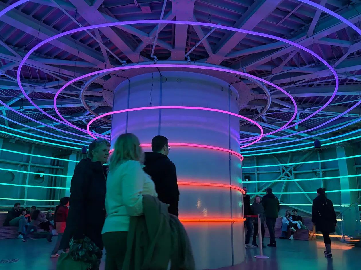 Raum in einer Kugel des Atomiums mit einer weißen Mittelsäule, um die leuchtende Ringe in Lila, Rosa und Türkis hängen, dazwischen Besucher