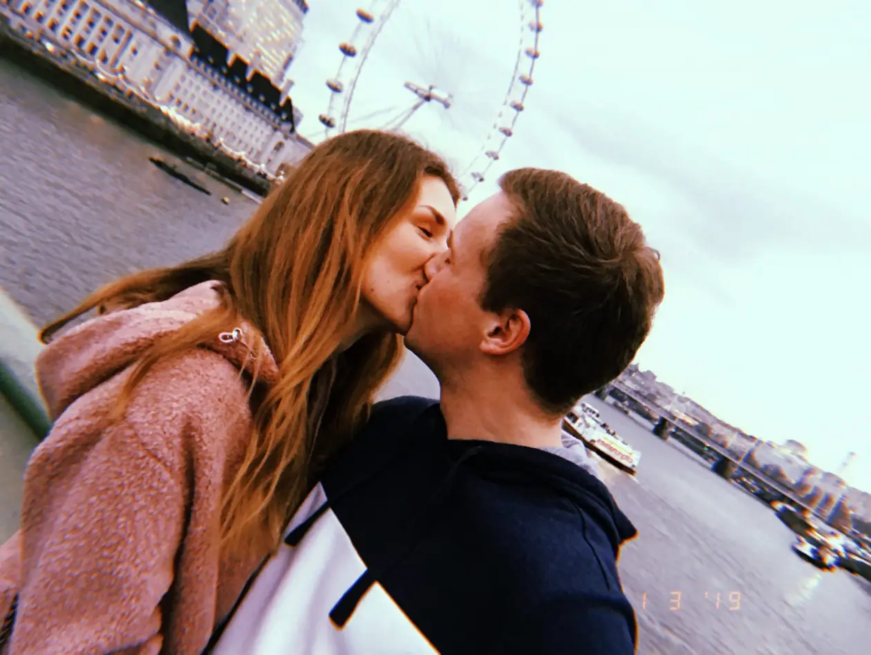 Max und Rebecca küssen sich am Geländer der Westminster Bridge, schräg im Bild dahinter die Themse und das London Eye