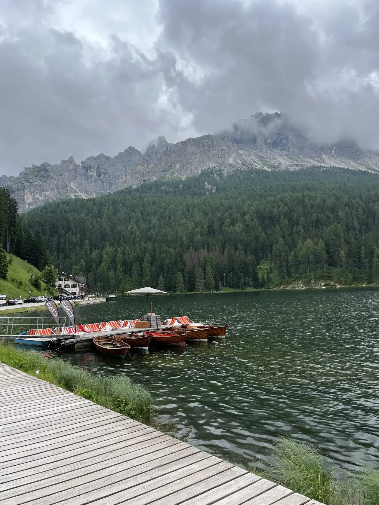 Holzsteg am Ufer des Lago di Misurina, am Anleger Ruder- und Tretboote, gegenüber bewaldete Hänge und ein Felsmassiv in tief hängenden Wolken