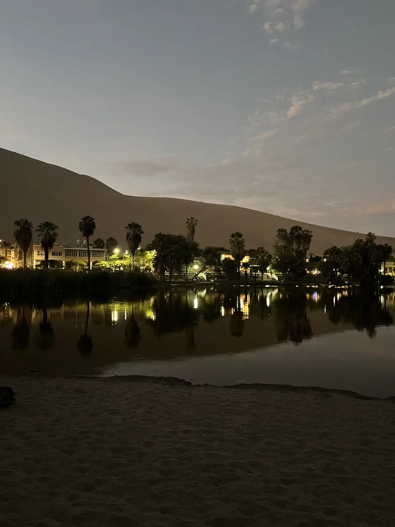 Die Lagune in der Dämmerung, Lichter und Palmen spiegeln sich im Wasser, dahinter der dunkle Umriss einer Düne