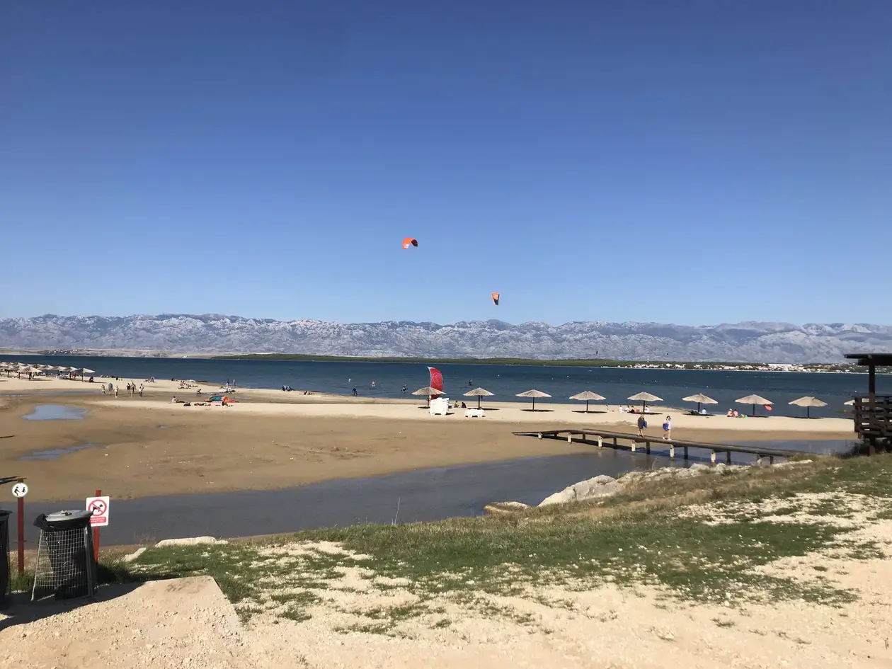 Sandstrand an einer flachen Lagune mit Strohschirmen und einem Holzsteg, über dem Wasser Kitesurfer, dahinter ein kahler Gebirgszug