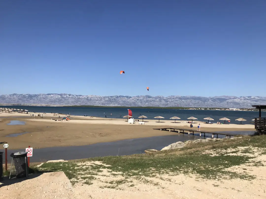 Sandstrand an einer flachen Lagune mit Strohschirmen und einem Holzsteg, über dem Wasser Kitesurfer, dahinter ein kahler Gebirgszug