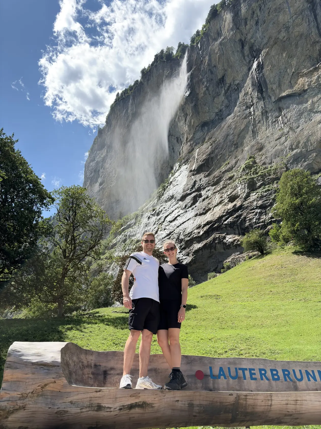 Max und Rebecca stehen auf einem Holzstamm mit der Aufschrift Lauterbrunnen, hinter ihnen ein Wasserfall, der über eine hohe Felswand stürzt