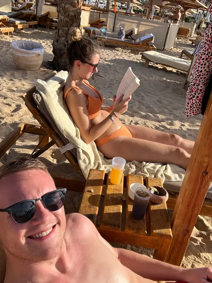 Rebecca liest auf einer Strandliege ein Buch, im Vordergrund grinst Max in die Kamera, zwischen den beiden ein Tisch mit Getränken
